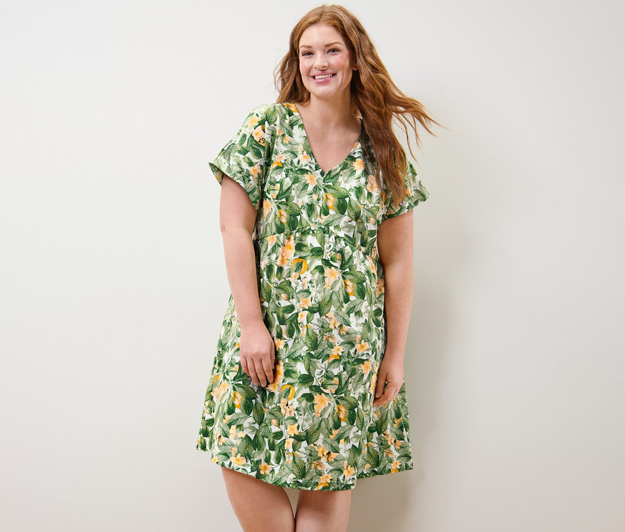 Žena s dlhými ryšavými vlasmi stojí a usmieva sa. Má na sebe džersejové šaty s potlačou.