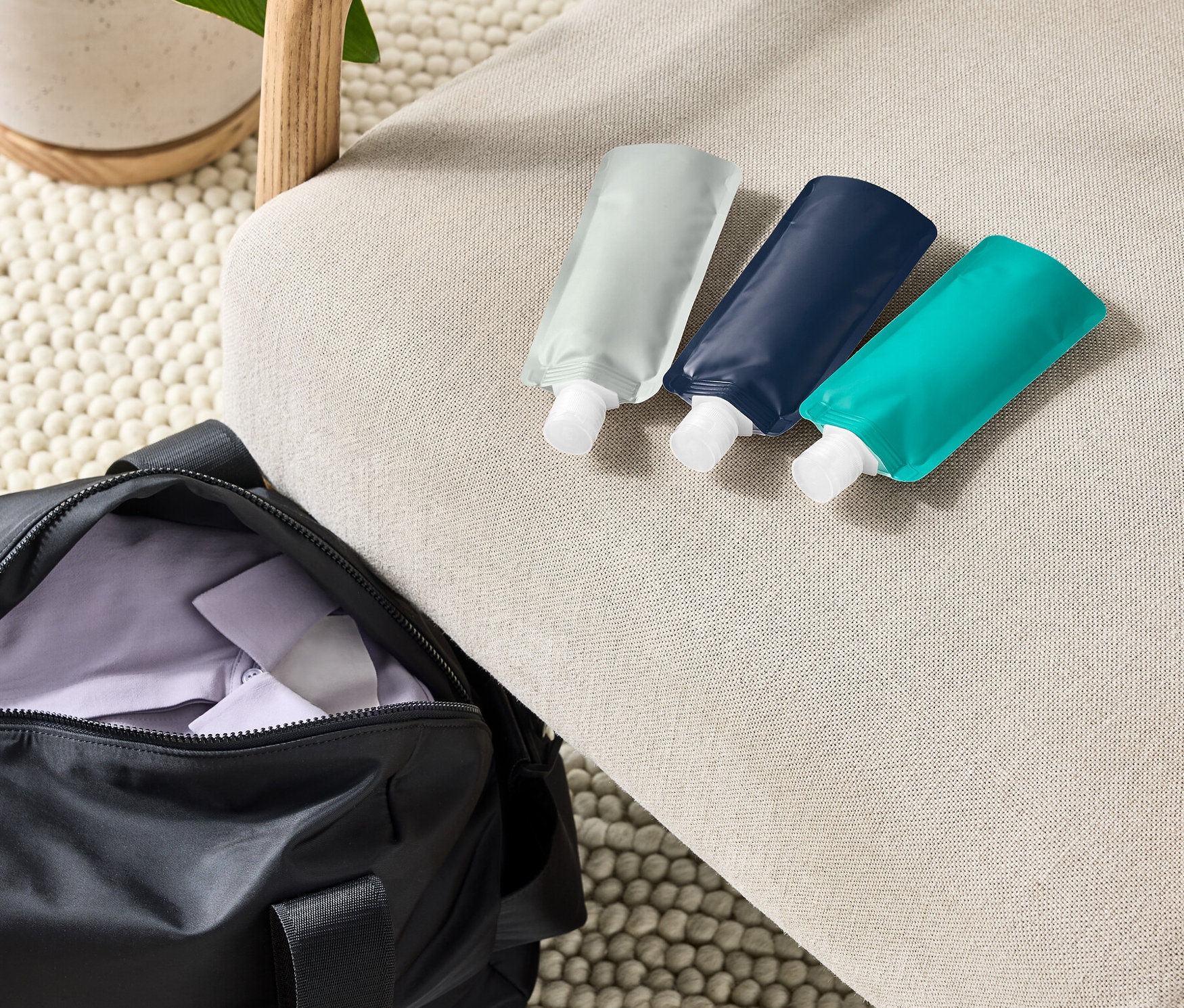 Tri opakovane naplniteľné cestovné fľaštičky ležia na béžovom kresle vedľa čiernej športovej tašky a rastliny v kvetináči.