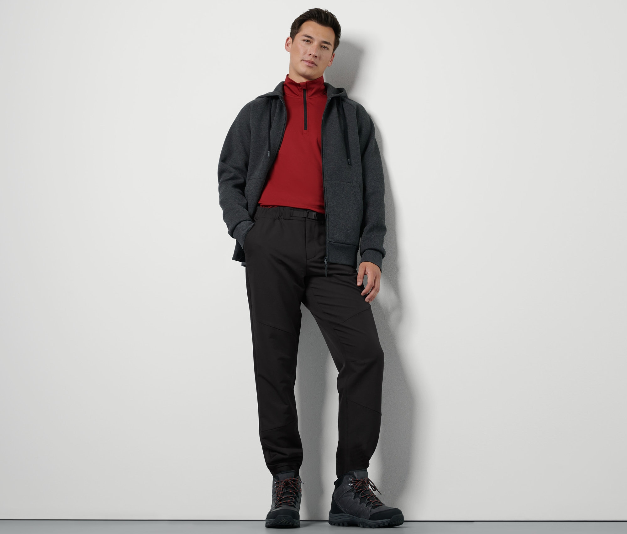 Muž stojí a má na sebe termonohavice s opaskom, mikinu na zips a turistické topánky.