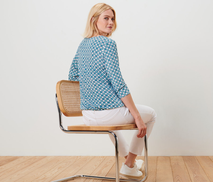 Žena sedí na stoličke a má na sebe blúzkové tričko s celoplošnou potlačou.