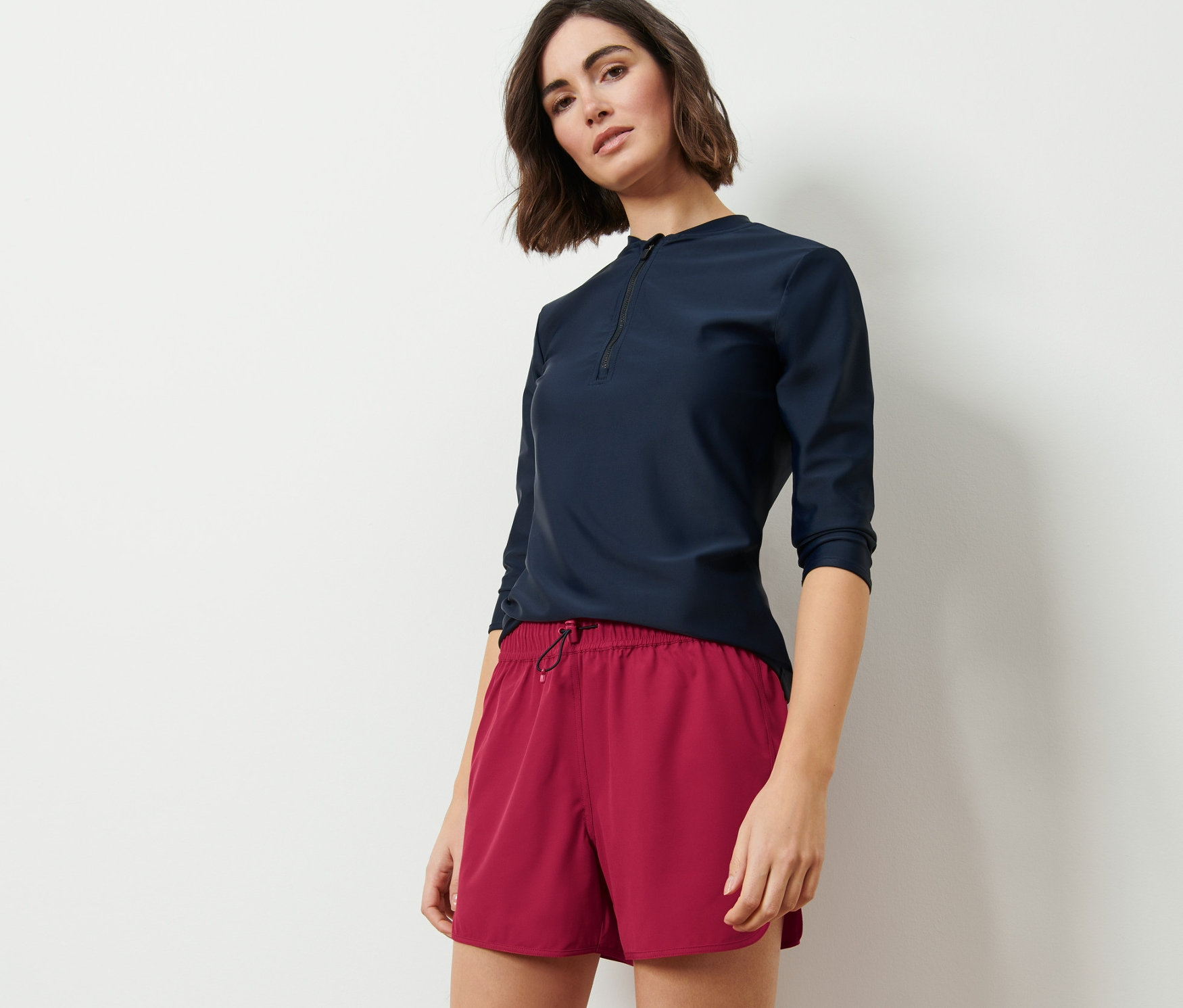 Žena pózuje v tmavomodrom tope s trojštvrťovými rukávmi a vínových šortkách na kúpanie.