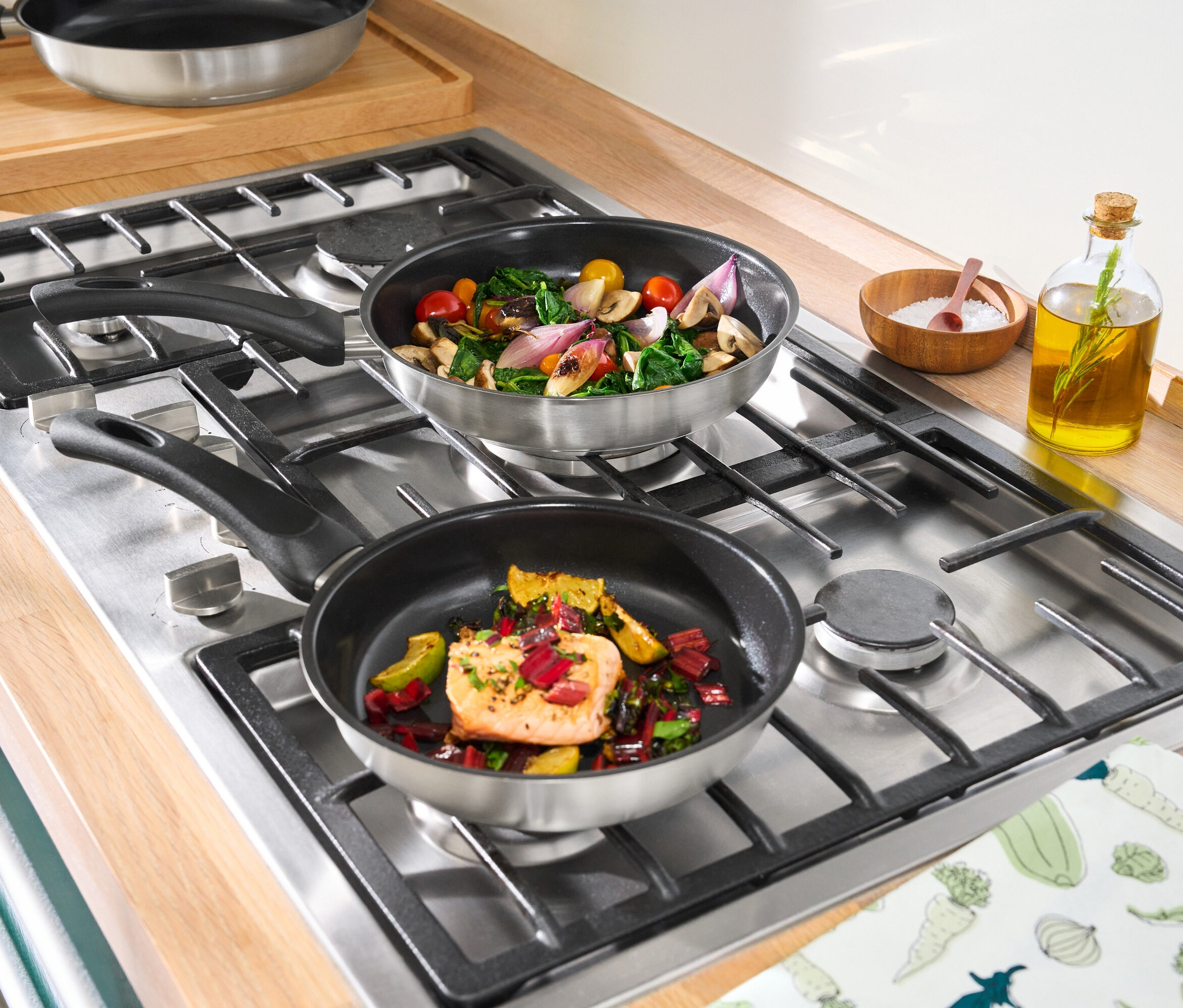 Dve panvice z ušľachtilej ocele s keramickým povrchom na sporáku. V jednej panvici je losos so zeleninou, v druhej zelenina.