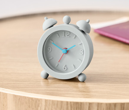 Sivý cestovný budík na drevenom stolíku vedľa pasu.