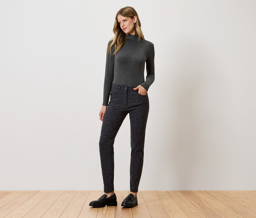 Žena stojí a pózuje v sivom roláku, vzorovaných nohaviciach a čiernych topánkach.