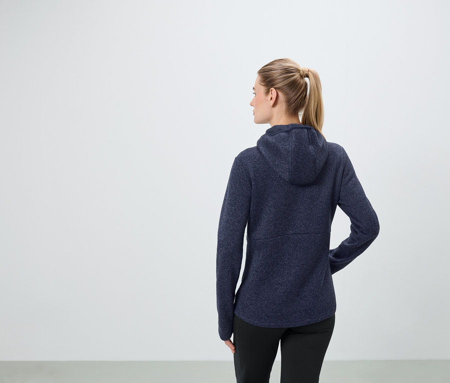 Žena s blond vlasmi zopnutými do copu má na sebe tmavomodrú mikinu s kapucňou a čierne nohavice.