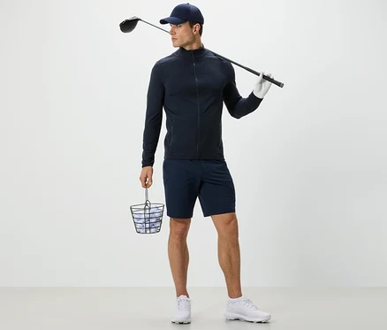 Muž v tmavomodrej funkčnej bunde drží kôš s golfovými loptičkami a golfovú palicu.