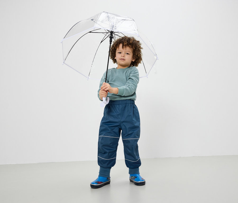 Chlapec drží priehľadný dáždnik, má na sebe modré nohavice a gumáky.