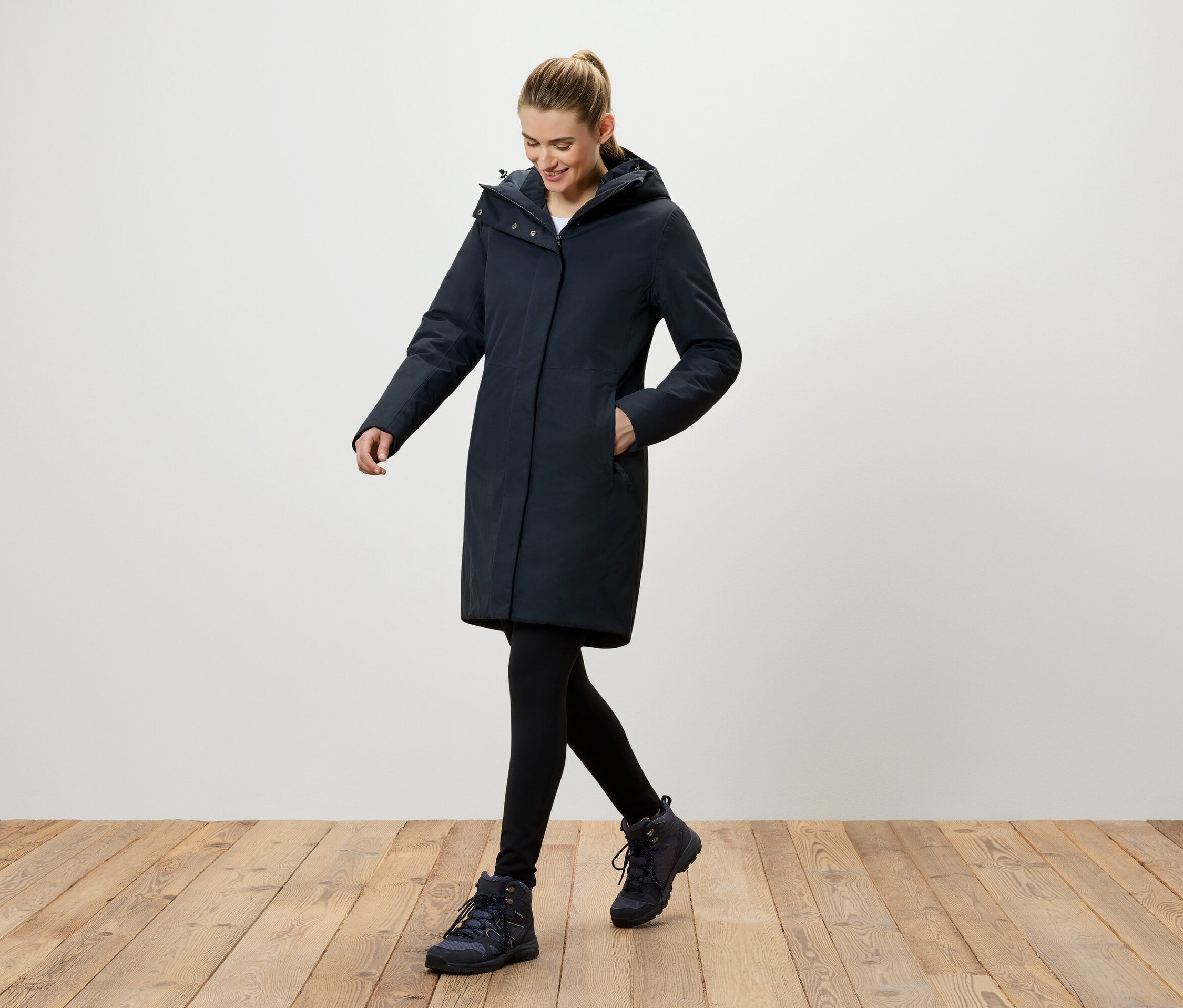 Žena kráča po drevenej podlahe a má na sebe tmavomodrý kabát s kapucňou, čierne legíny a tmavomodré topánky.