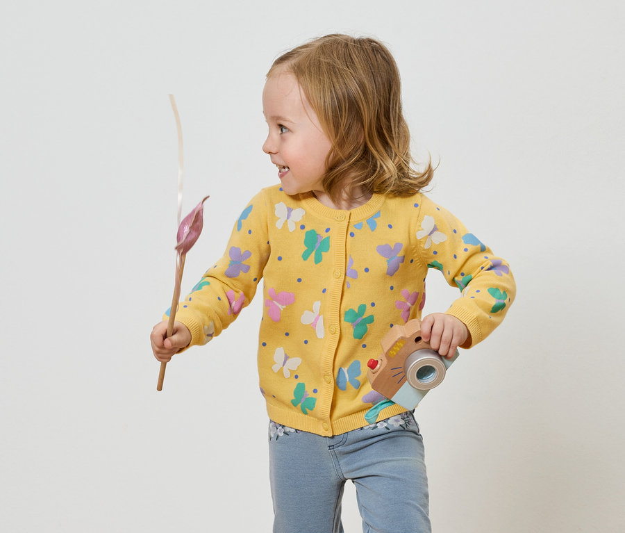 Dievča v žltom svetri s motýľmi drží hračkársky fotoaparát a kvetinu.