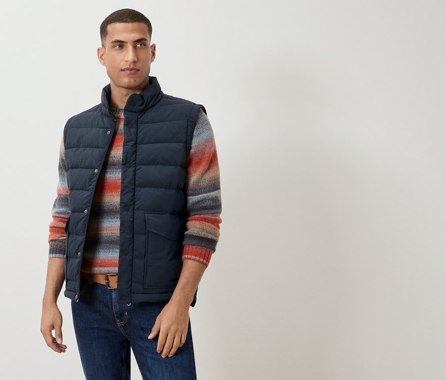 Muž má na sebe tmavomodrú vestu cez pruhovaný sveter, hnedý opasok a džínsy.