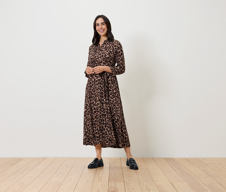 Žena pózuje vo vzorovaných šatách s dlhým rukávom a čiernych topánkach.