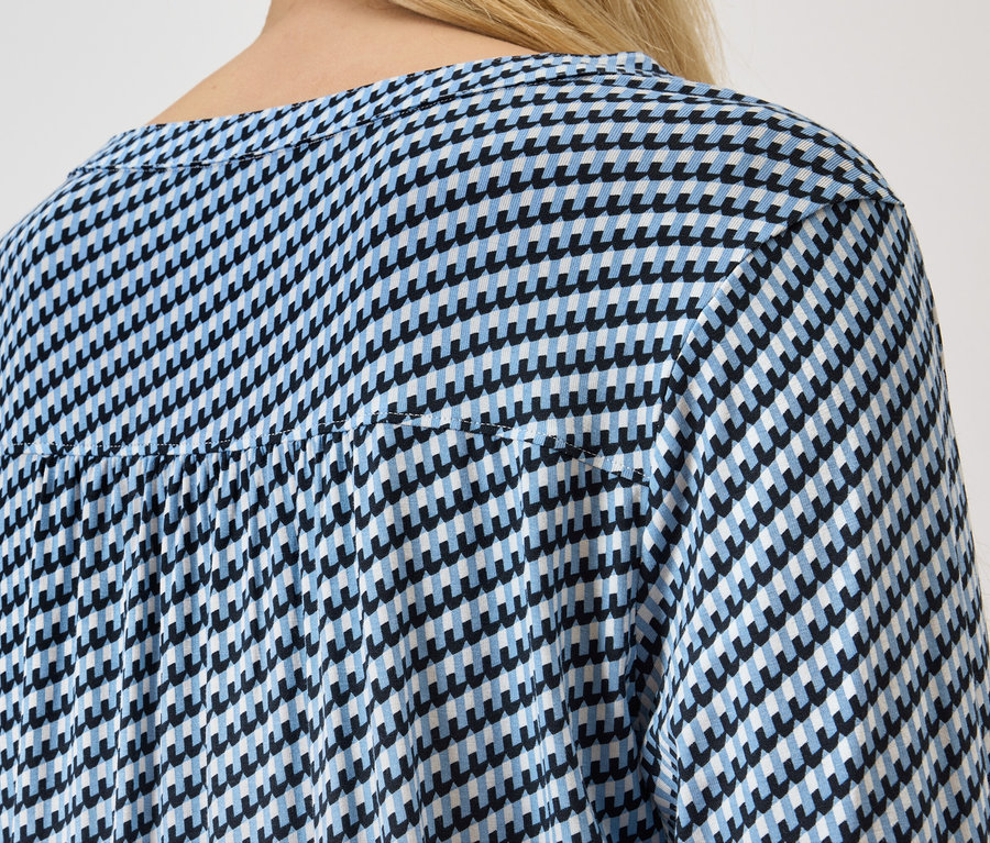 Pohľad zozadu na ženu s blond vlasmi v modro-bielej blúzke s kohútím vzorom.