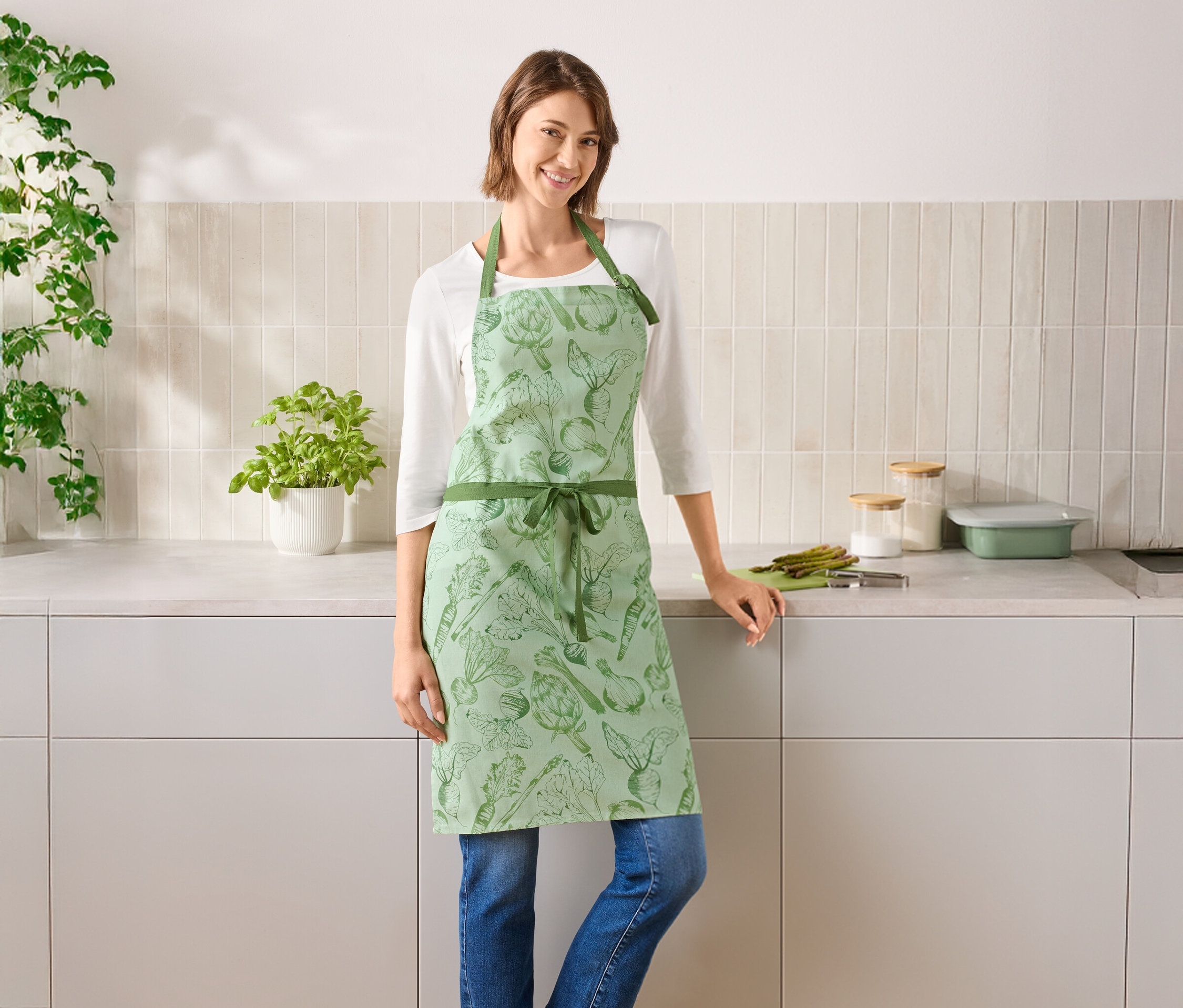 Žena stojí v kuchyni a má na sebe zelenú zásteru so zeleninou.