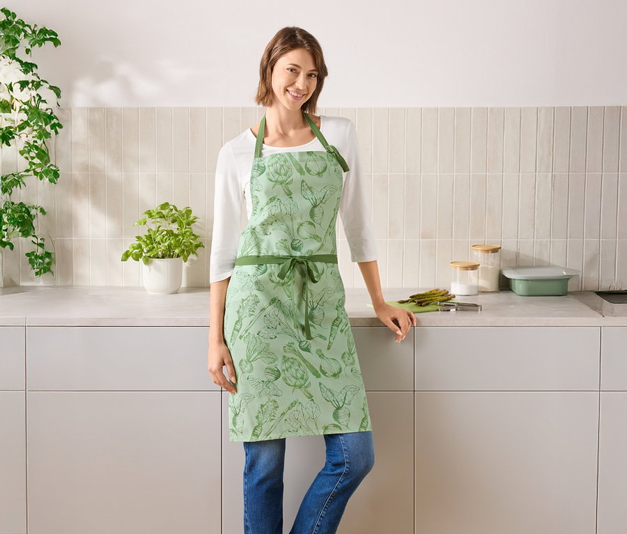 Žena stojí v kuchyni a má na sebe zelenú zásteru so zeleninou.