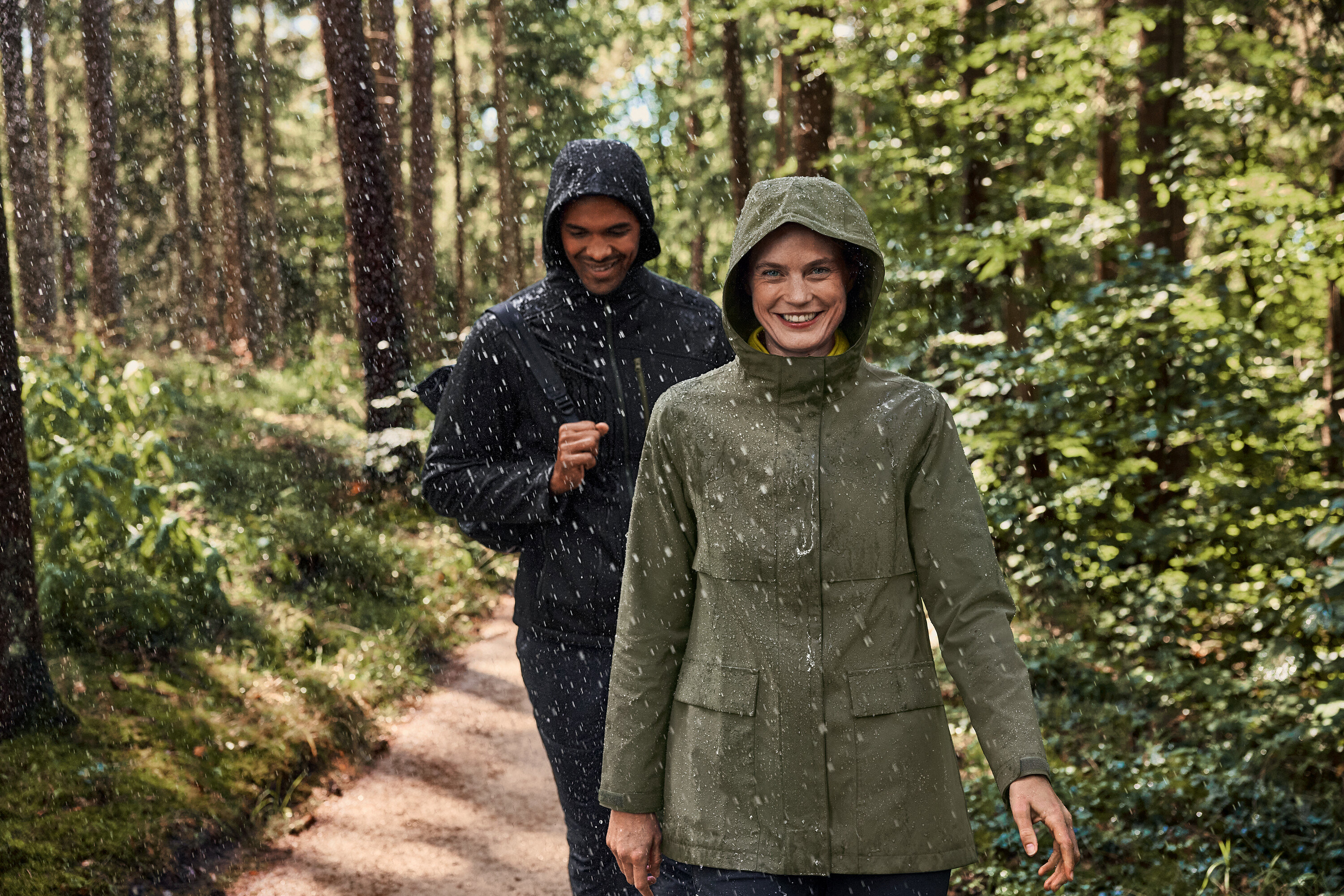 Dvaja ľudia kráčajú v daždi cez les. Žena má na sebe flísový sveter kaki-antracitový.