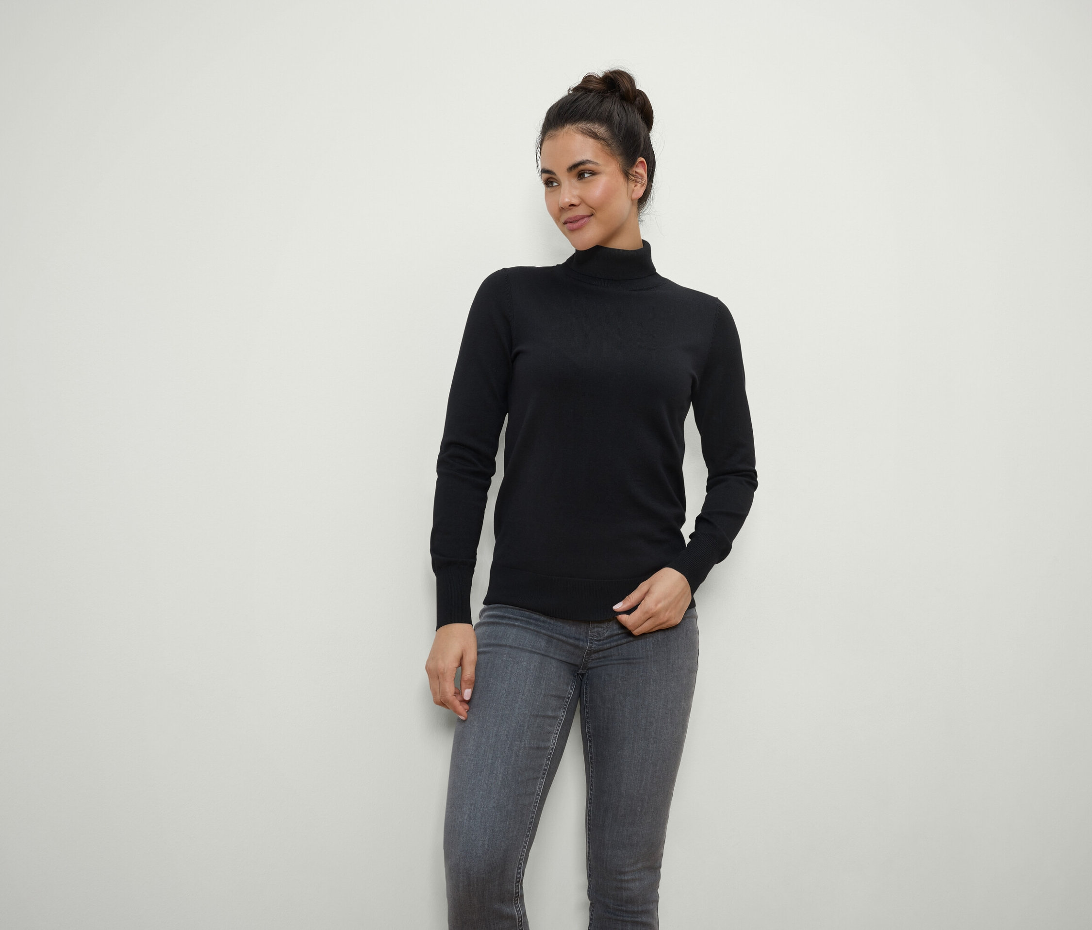 Žena s hnedými vlasmi stiahnutými do drdola pózuje v čiernom roláku a sivých džínsoch.