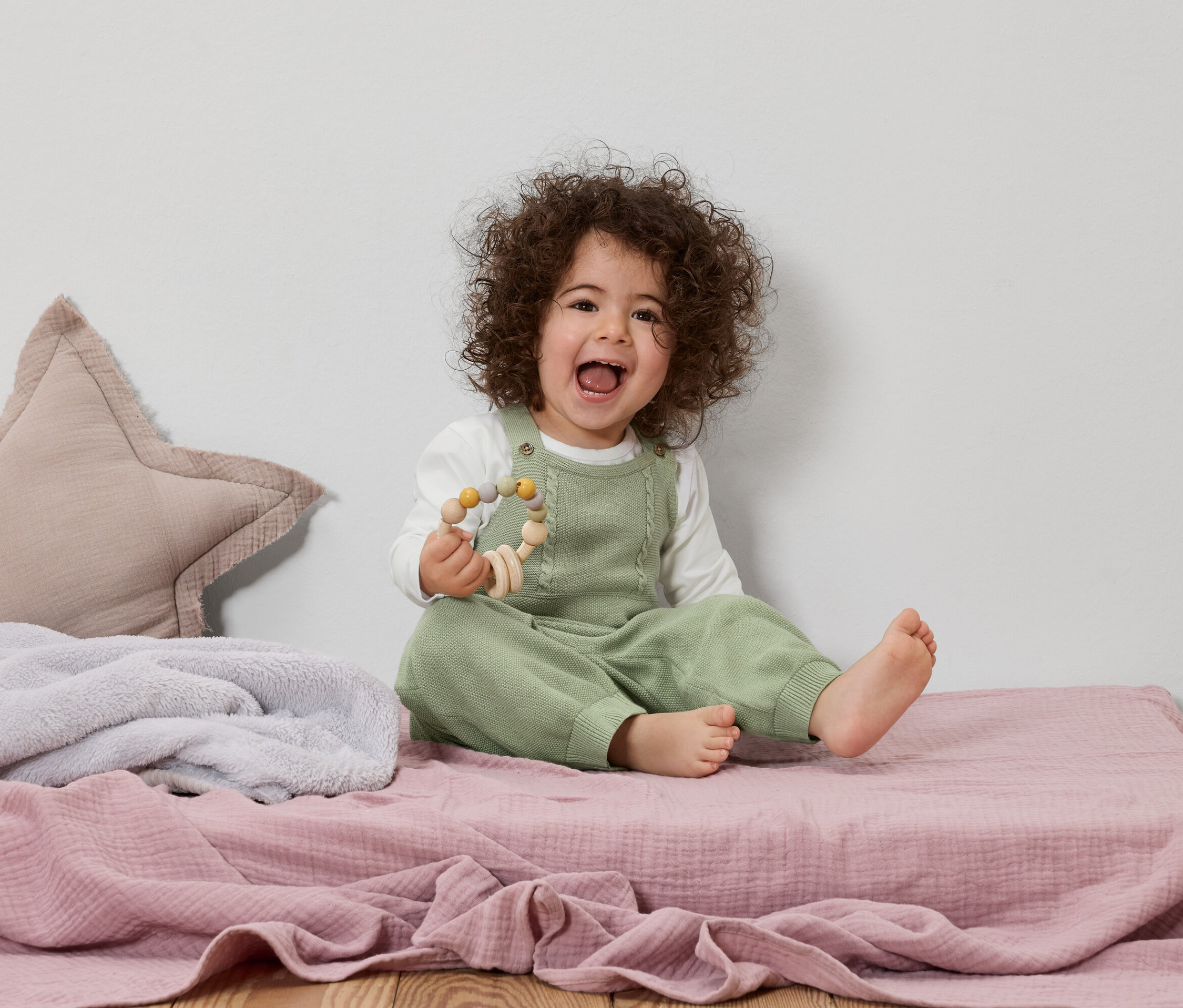 Smejúce sa dieťa s kučeravými vlasmi sedí na ružovej hracej deke a drží drevenú hračku.