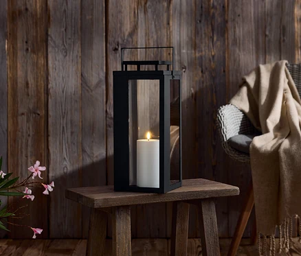 Čierna lampa so sviečkou na drevenom stole. V pozadí drevená stena a stolička s dekou.