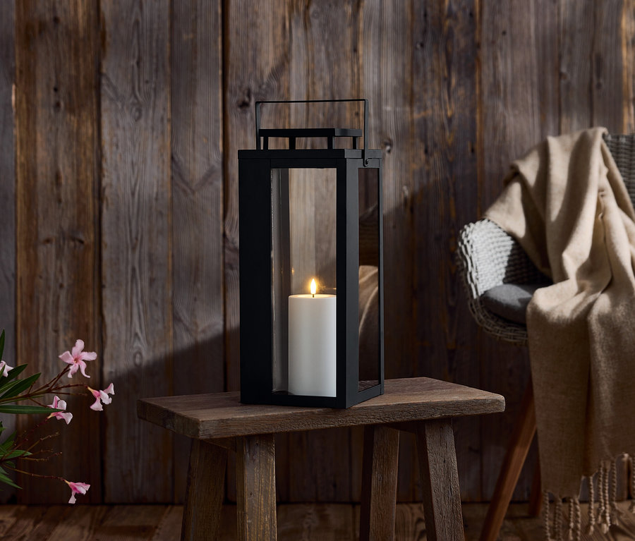 Čierna lampa so sviečkou na drevenom stole. V pozadí drevená stena a stolička s dekou.