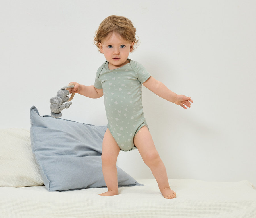 Batoľa v zelenom body s potlačou srdiečok stojí na posteli a drží sivú hračku slona.
