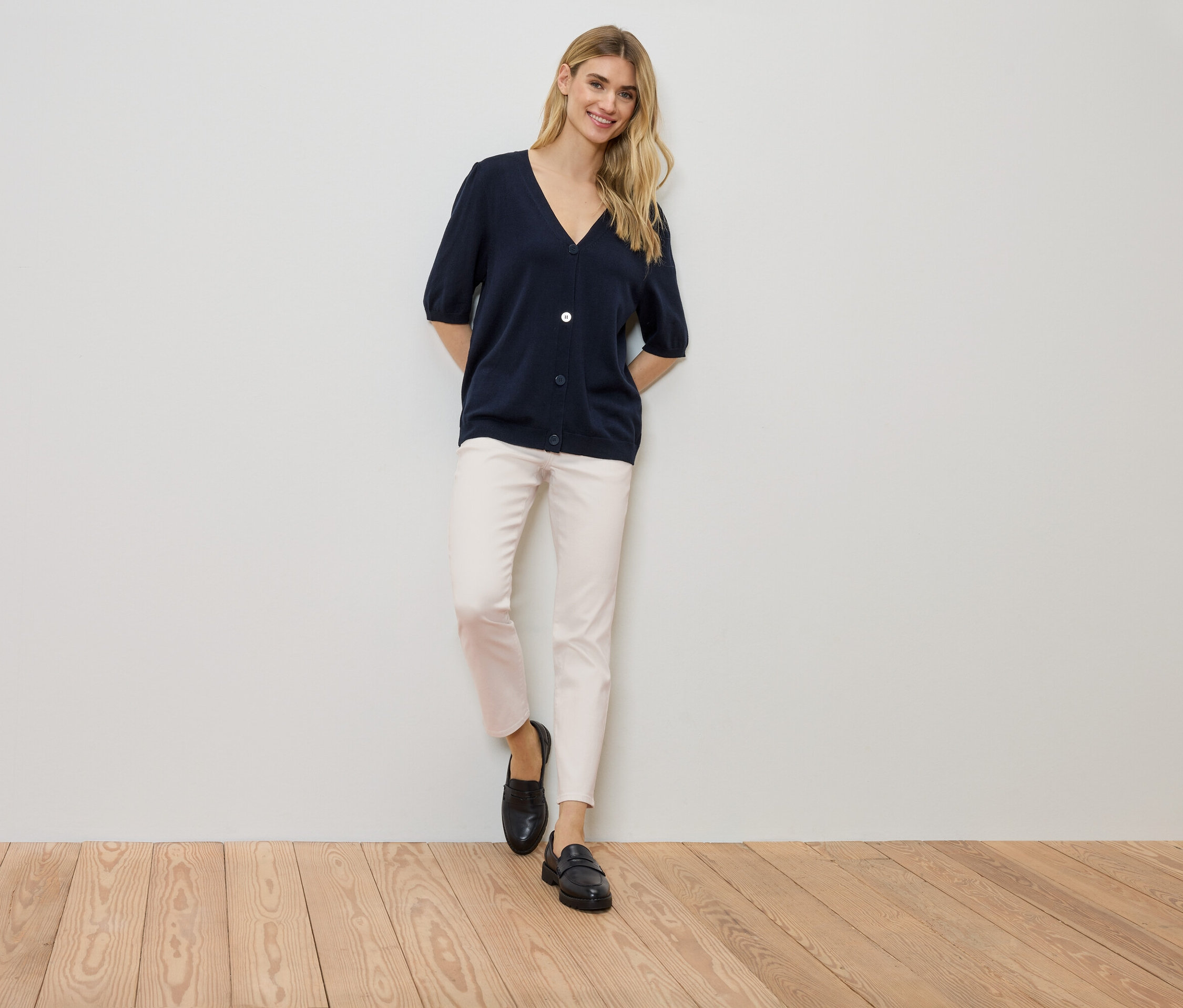 Žena stojí pri stene, má na sebe tmavomodrý kardigán, svetlé nohavice a čierne topánky.