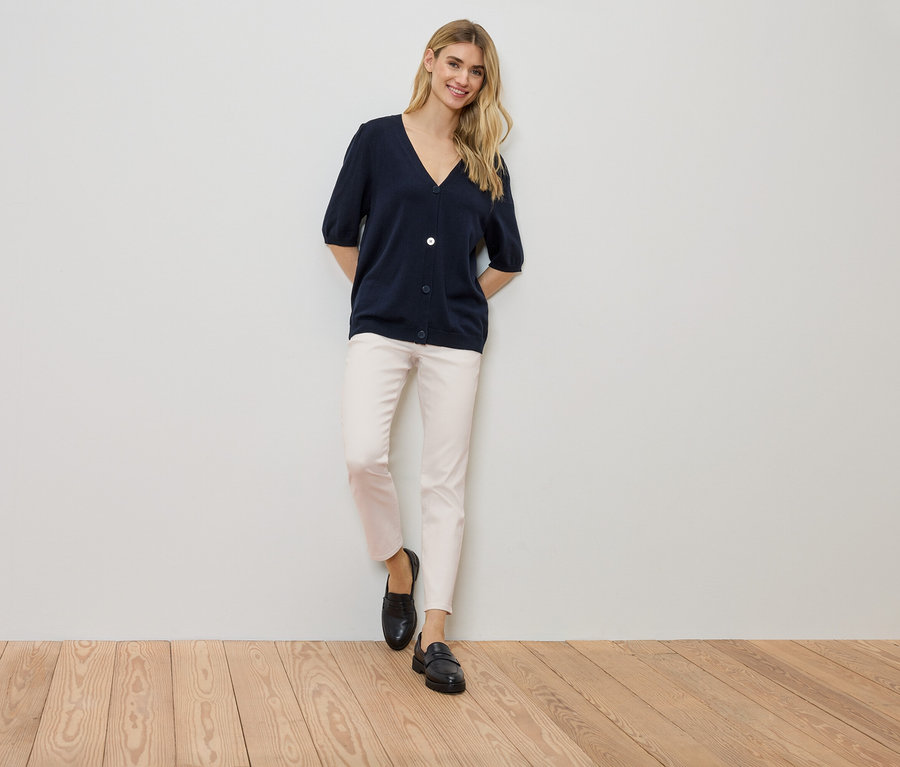 Žena stojí pri stene, má na sebe tmavomodrý kardigán, svetlé nohavice a čierne topánky.