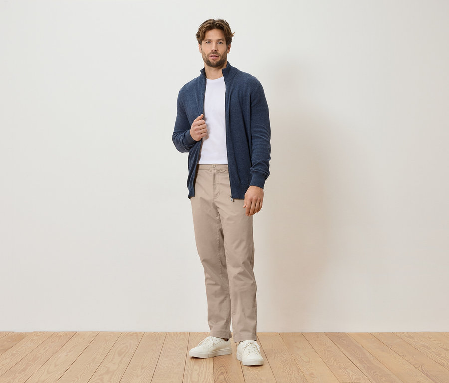 Muž stojí pred bielou stenou, má na sebe tmavomodrý kardigán, biele tričko, béžové nohavice a biele tenisky.