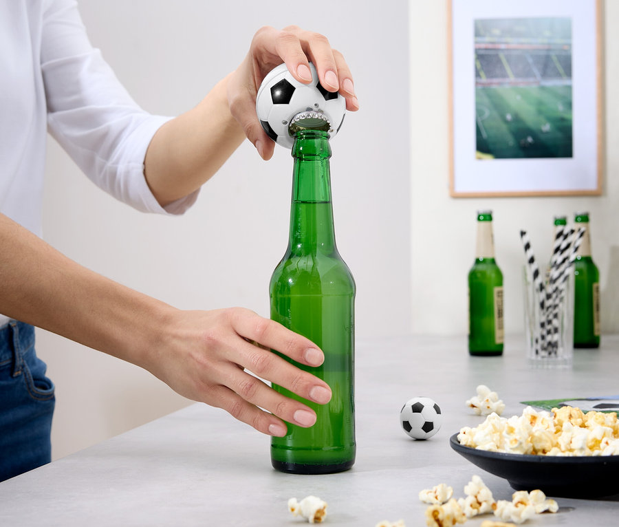 Osoba otvára zelenú fľašu piva otváračom na fľaše v tvare futbalovej lopty. Na stole sú popcorn a iné fľaše.