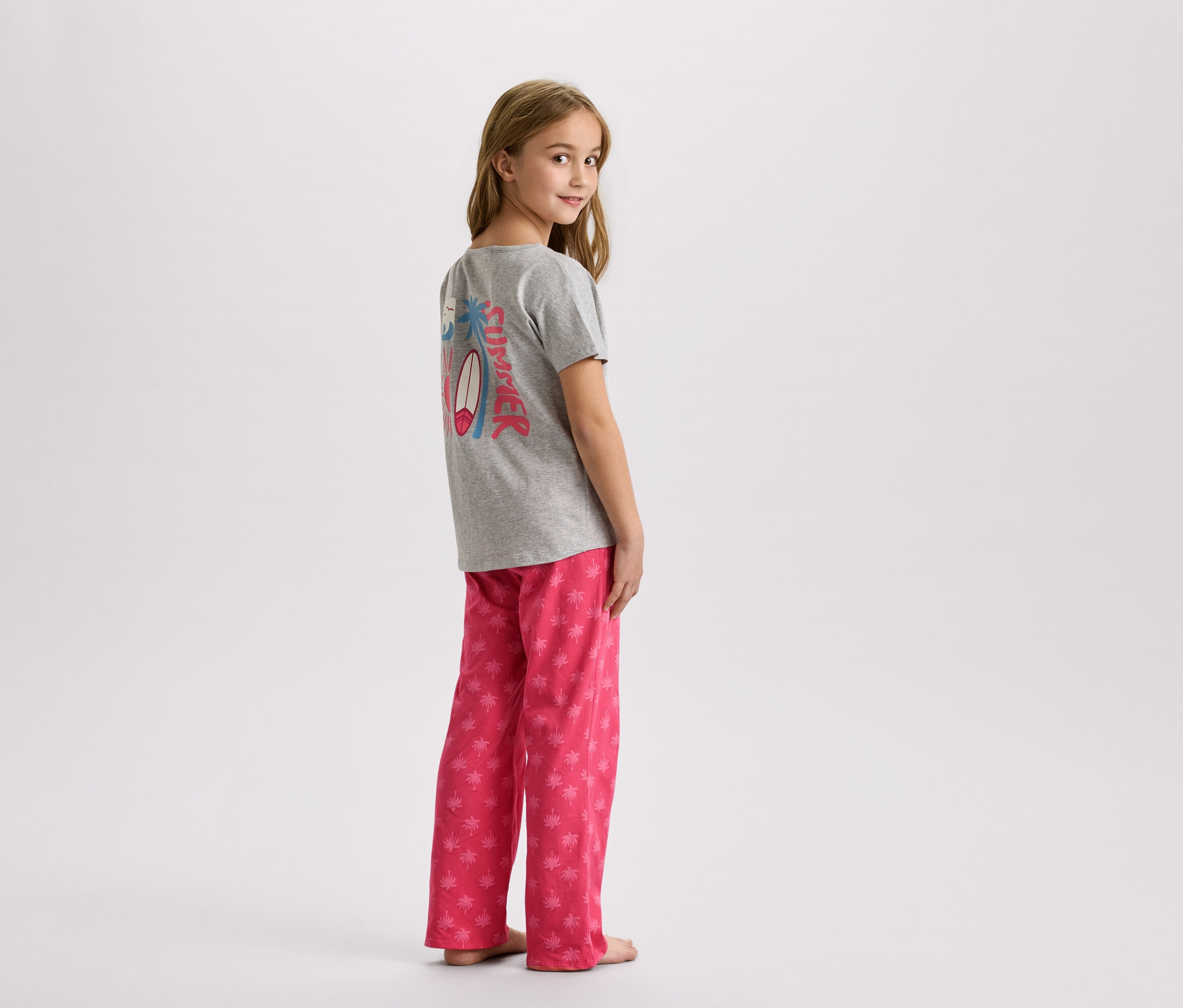 Dievča stojí v sivom tričku s letným dizajnom a ružových nohaviciach s palmami.