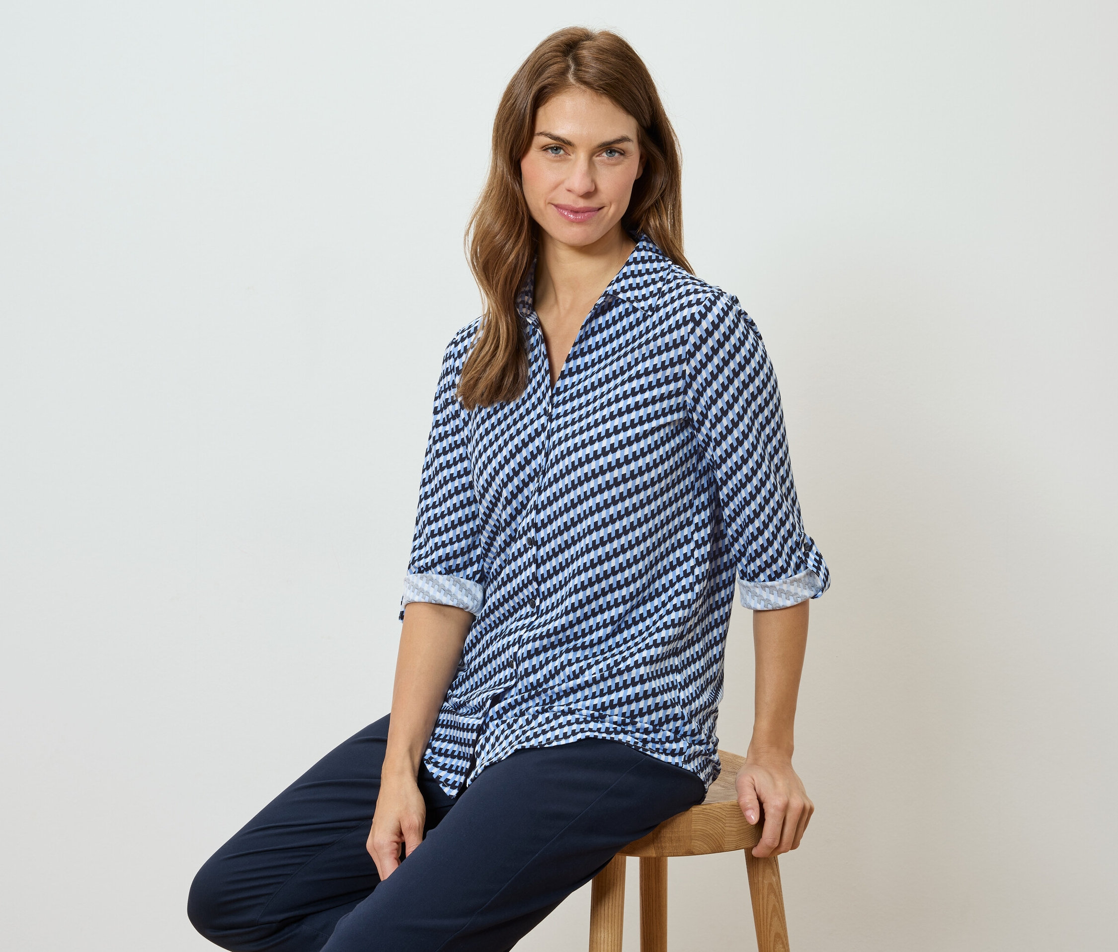 Žena sedí na stoličke a má na sebe modro-bielu blúzku so vzorom a tmavé nohavice.