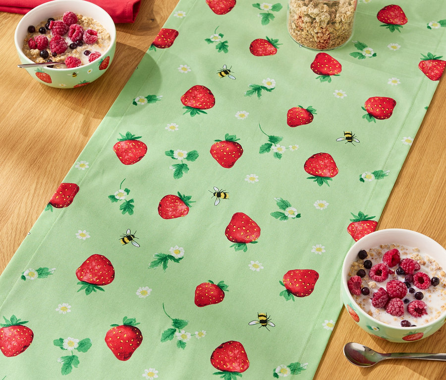 Na drevenom stole leží behúň so vzorom jahôd, na ňom misky s cereáliami, pohár müsli a fľaša mlieka.