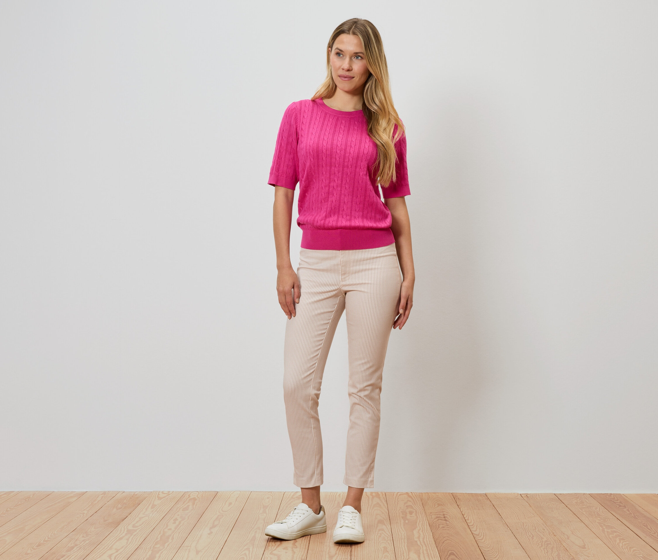 Žena stojí pred stenou, má na sebe ružový sveter, pásikavé nohavice a biele topánky.