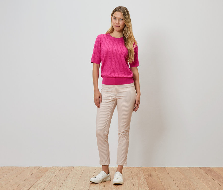 Žena stojí pred stenou, má na sebe ružový sveter, pásikavé nohavice a biele topánky.