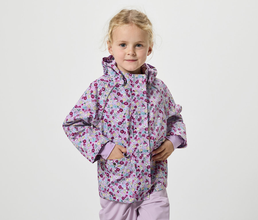 Dievča s blond vlasmi a fialovou kvetinovou bundou stojí na bielom pozadí.