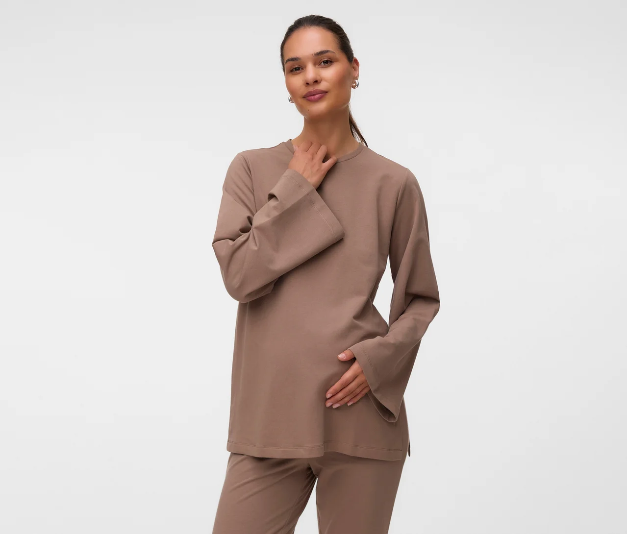 Tehotná žena má na sebe svetlohnedý MAMALICIOUS Sweat Top »MLSINE LS« a zladené nohavice, pravú ruku má na krku a ľavú na bruchu.