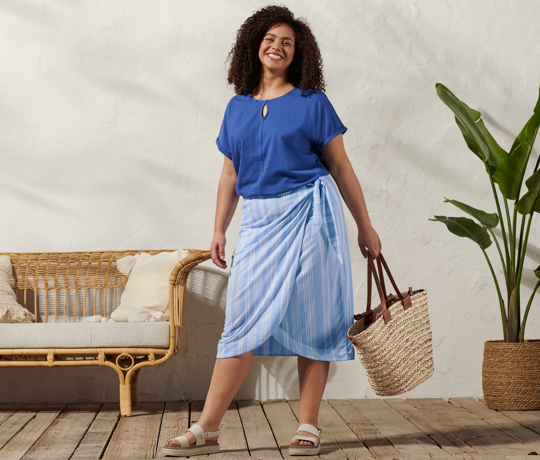 Žena má na sebe tričko z ľanovej zmesi, prúžkovanú zavinovaciu sukňu, kožené sandále na platforme a košíkovú tašku.