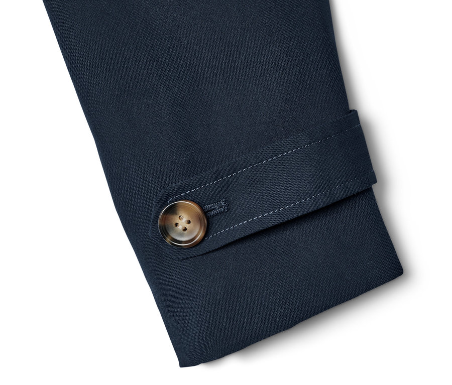 Detail rukávu námornícky modrého trenčkotu s hnedým gombíkom.