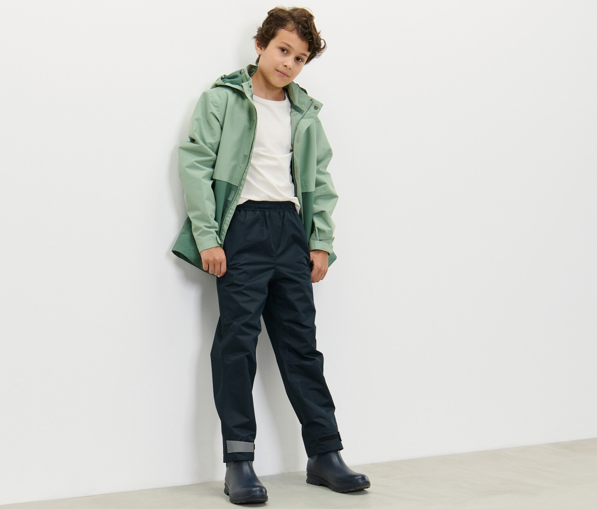 Chlapec v zelenej bunde a tmavomodrých detských nohaviciach do dažďa stojí opretý o bielu stenu.