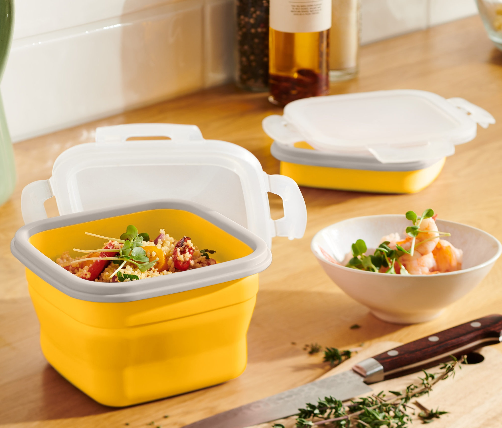 Žltá skladacia dóza na potraviny s obsahom stojí na drevenom stole vedľa zeleného džbánu a misy s krevetami.