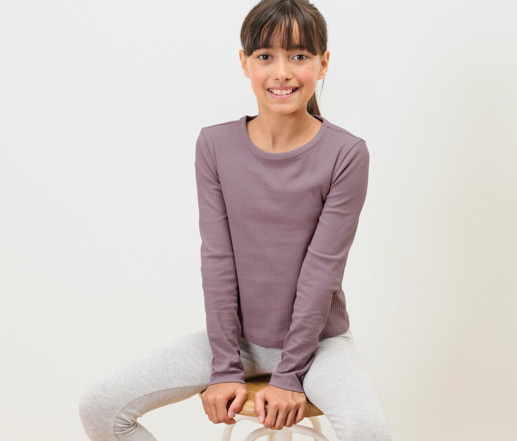 Dievča sedí na stoličke a má na sebe detské tričko s dlhým rukávom s rebrovanou štruktúrou.