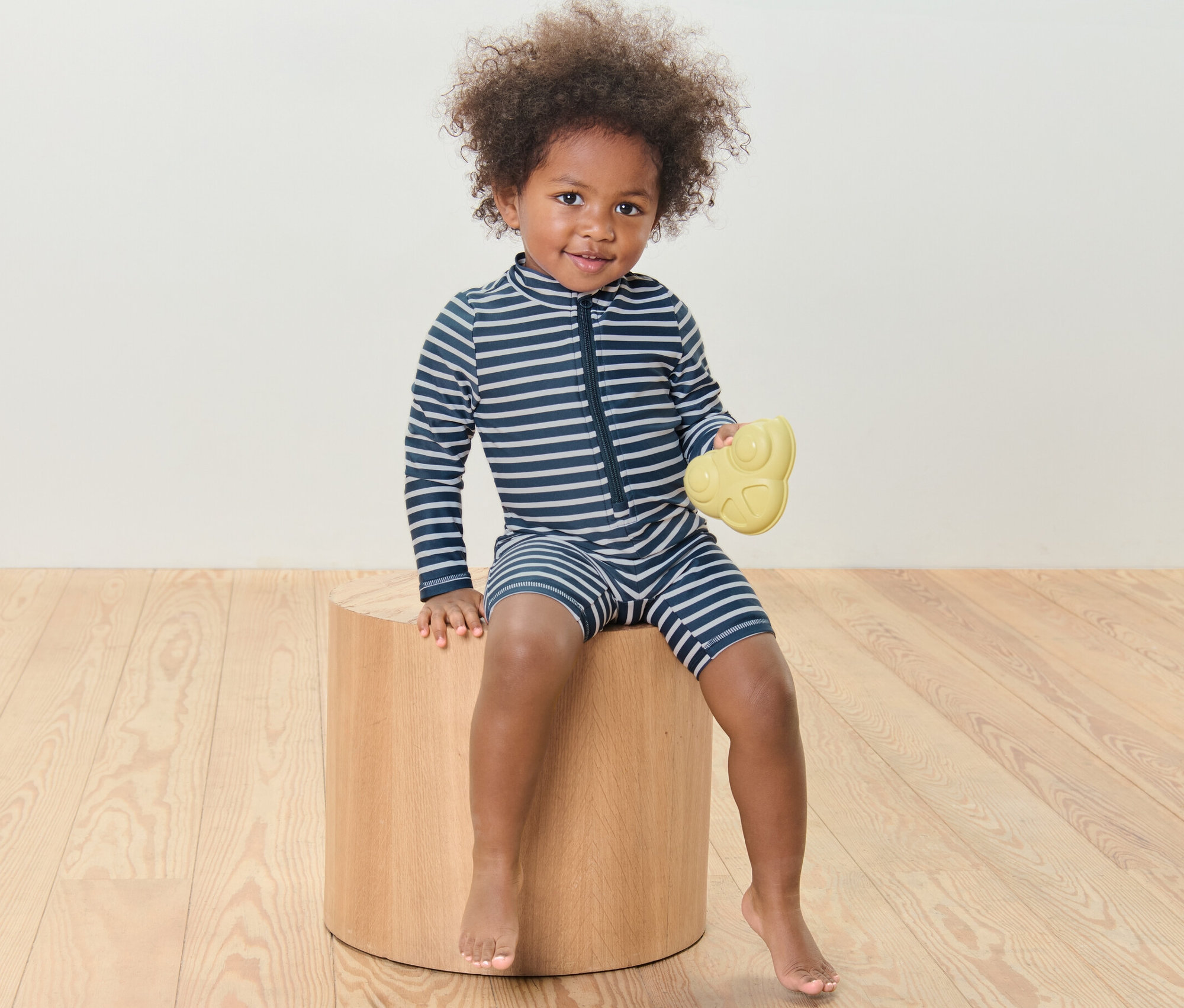 Dieťa sedí na drevenej stoličke a drží žltú hračku, má na sebe detský overal s UV ochranným faktorom 80, pásiky.