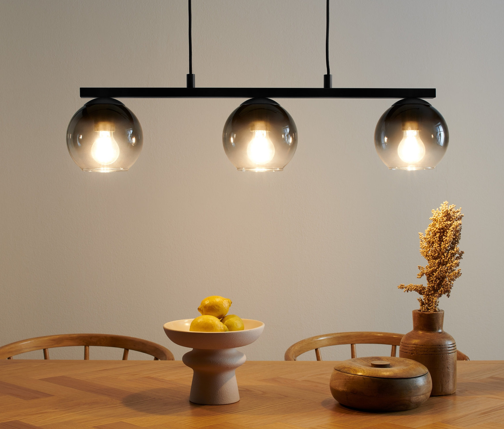 Závesné svietidlo s tienidlami z dymového skla nad dreveným stolom. Na stole leží misa s ovocím a drevená dóza.