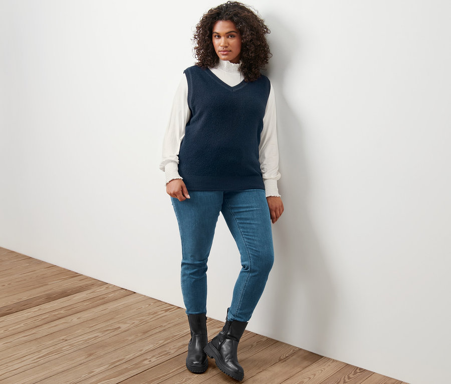 Žena pózuje v pletenej veste so vzhľadom buklé, krémovom blúzkovom tričku so stojačikom a tregínsoch »Fit Nelly«.