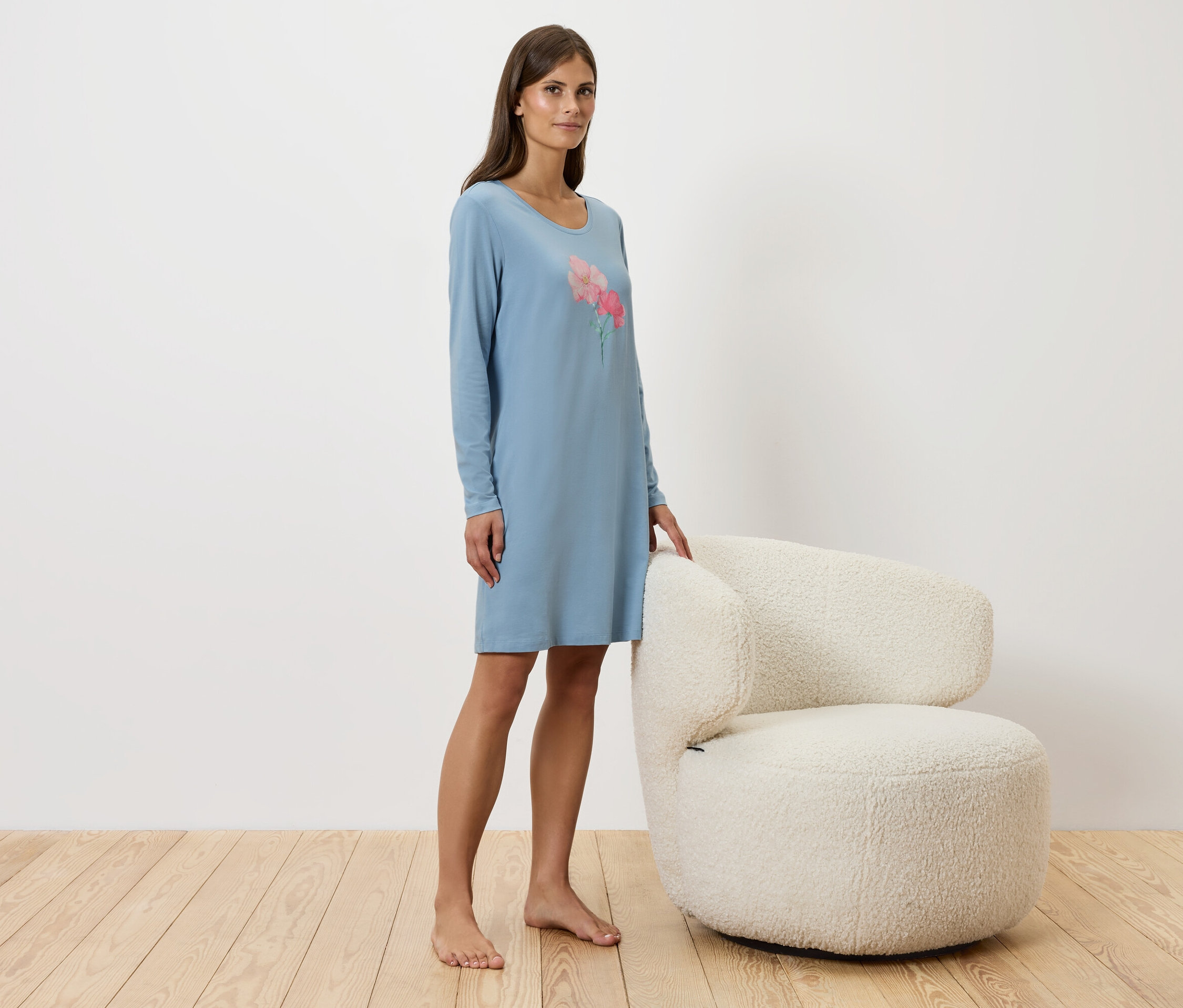 Žena stojí vedľa bieleho kresla a má na sebe modré nočné šaty.