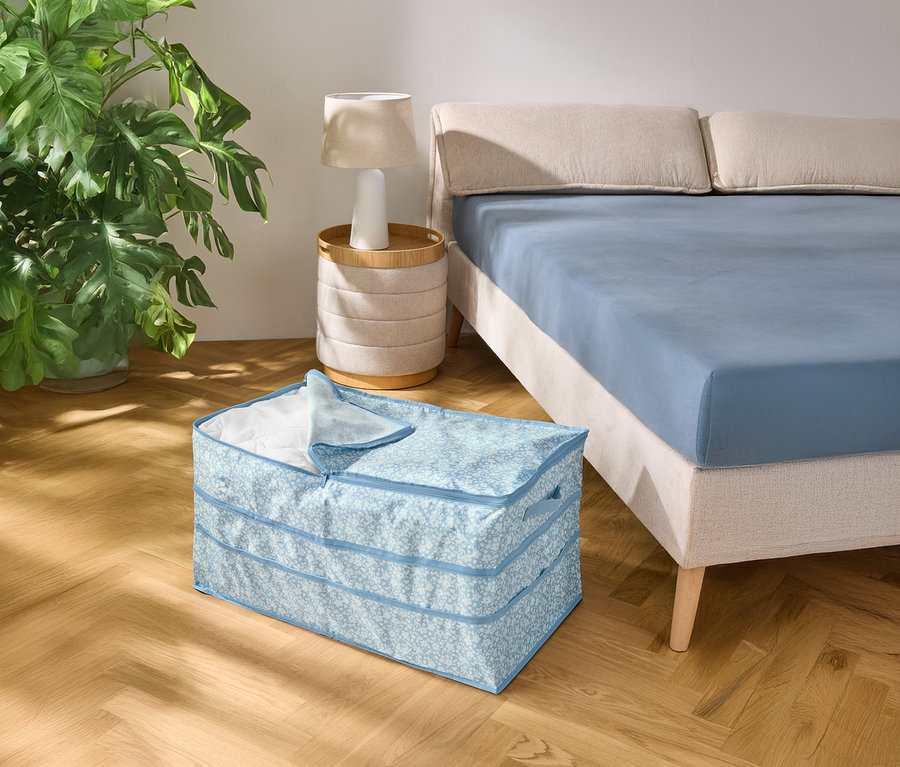 Na drevenej podlahe stojí svetlomodrá vzorovaná úložná škatuľa vedľa postele s modrou plachtou.