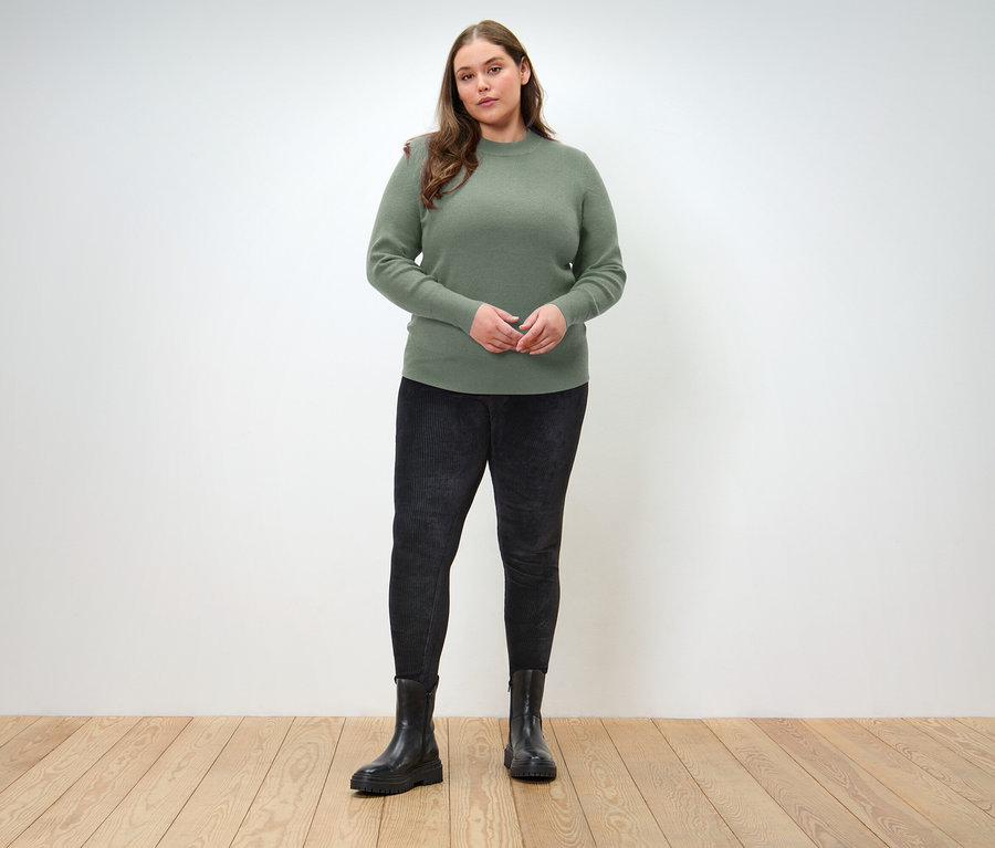 Žena stojí na drevenej podlahe a má na sebe zelený sveter, čierne džínsy a čierne topánky.