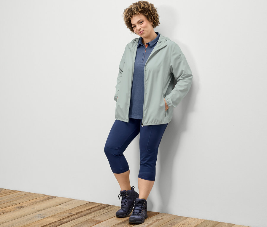 Žena opretá o stenu má na sebe 3/4 trekingové legínsy, tmavomodrú funkčnú polokošeľu a sivú skladaciu funkčnú bundu.