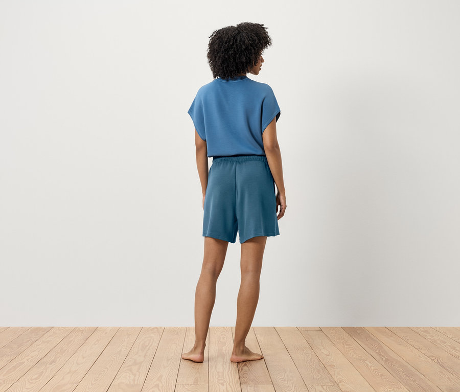 Žena v modrom tričku a šortkách stojí chrbtom k fotoaparátu.