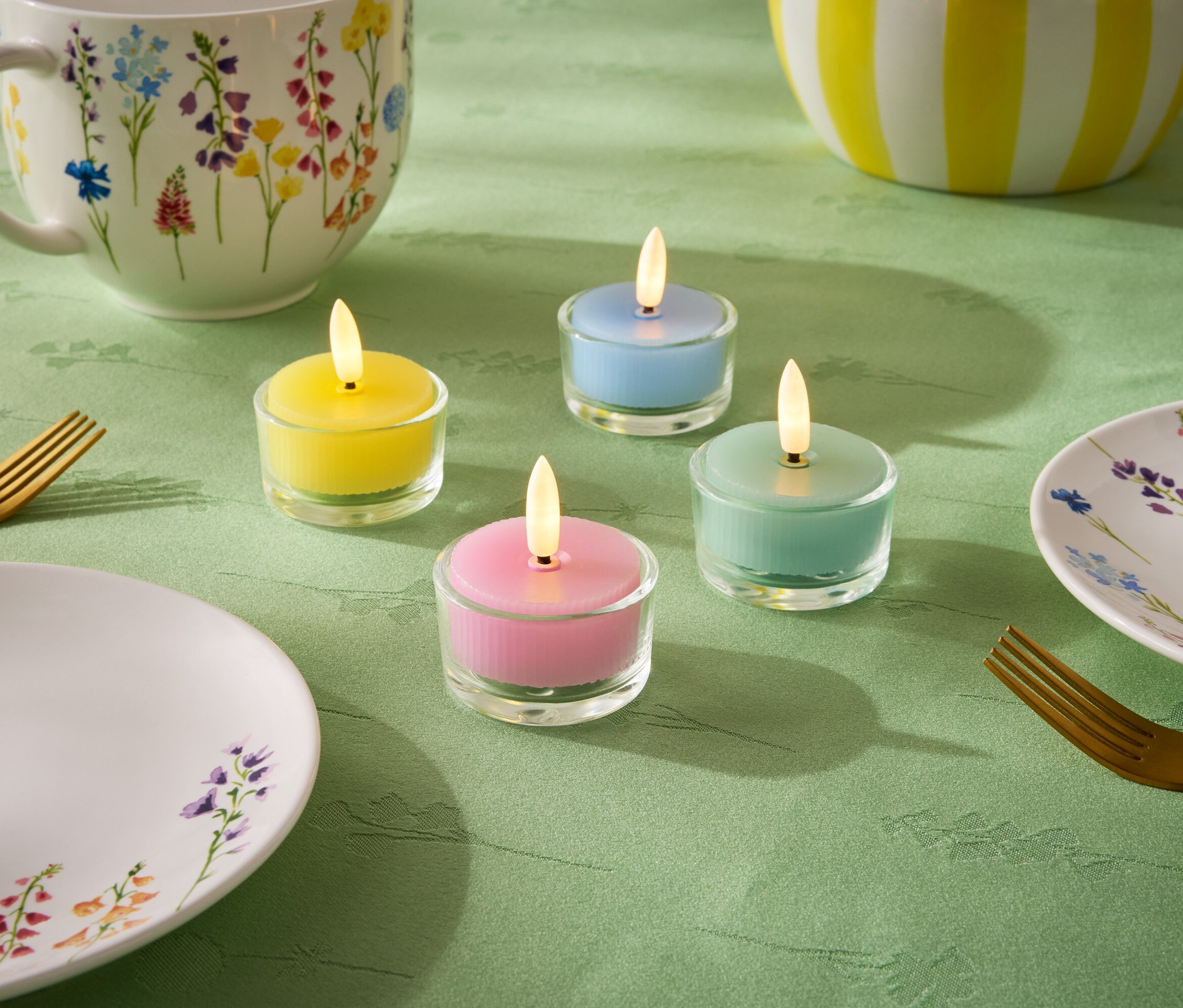 Na zelenom obruse sú štyri malé sviečky v pastelových farbách: žltá, modrá, zelená a ružová. Ďalej je tam tanier, šálka a misa s kvetinovým vzorom a dve zlaté vidličky.