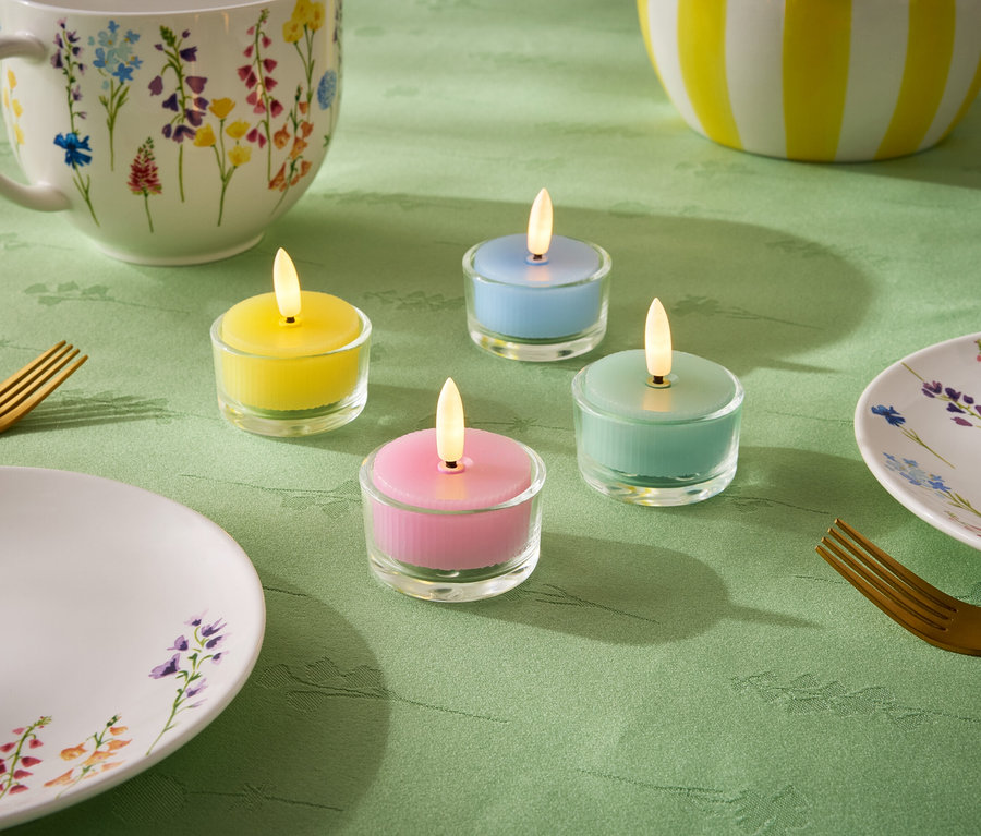 Na zelenom obruse sú štyri malé sviečky v pastelových farbách: žltá, modrá, zelená a ružová. Ďalej je tam tanier, šálka a misa s kvetinovým vzorom a dve zlaté vidličky.